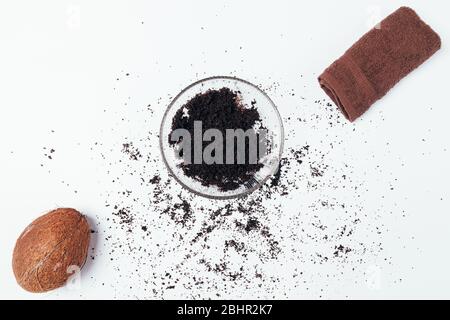 Scrub corpo caffè con olio di cocco in una ciotola di vetro accanto a un asciugamano marrone su un tavolo bianco, piatto. Foto Stock