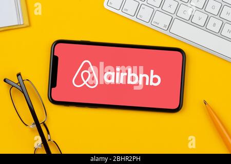 Uno smartphone con il logo Airbnb è posizionato su uno sfondo giallo insieme a tastiera, occhiali, penna e libro (solo per uso editoriale). Foto Stock