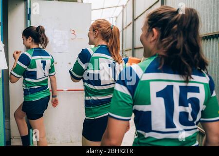 Vista posteriore di tre donne che indossano rugby blu, bianco e verde che camminano attraverso una porta in un edificio. Foto Stock