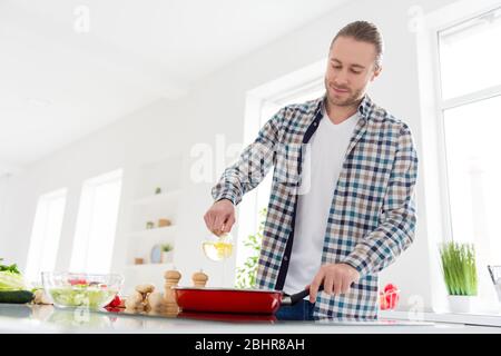 Foto di positivo bell'uomo cucinare gustoso delizioso pasto cena versando olio di girasole sulla padella indossare casual camicia a scacchi stand in cucina Foto Stock