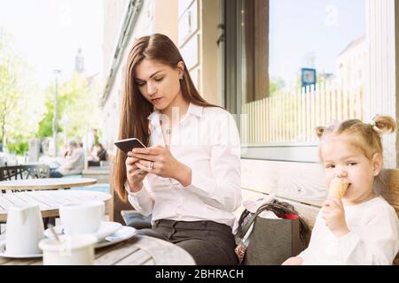 Una madre e una figlia seduti fuori da un bar, una madre sul suo cellulare, una figlia che mangia un gelato. Foto Stock