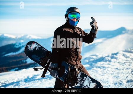 Una persona che indossa una tuta da sci nera, un casco e occhiali di protezione che tengono uno snowboard in piedi sulla cima di una montagna innevata. Foto Stock