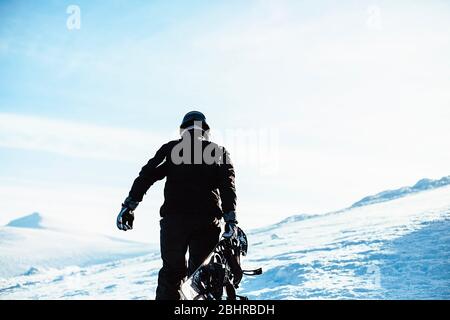 Una persona che indossa un costume da sci nero e un casco, con uno snowboard in piedi sulla cima di una montagna innevata. Foto Stock