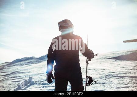 Silhouette di una persona che indossa una tuta da sci nera, casco e occhiali di protezione che tengono uno snowboard in piedi su una pista innevata. Foto Stock