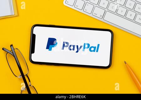 Uno smartphone con il logo PayPal è posizionato su uno sfondo giallo insieme a tastiera, occhiali, penna e libro (solo per uso editoriale). Foto Stock