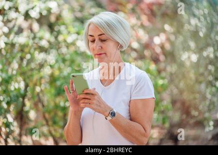 Donna anziana che si trova nel parco utilizzando smartphone e auricolari Foto Stock