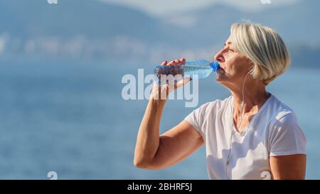 Donna matura bere acqua dopo jogging sullo sfondo il mare e cielo blu Foto Stock