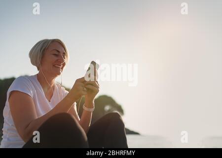 Donna matura seduta dopo jogging sulla spiaggia. Signora senior che usa smartphone e auricolari al tramonto. Corsa serale Foto Stock