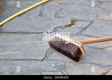 Vecchia scopa usata e tubo di irrigazione giallo che giace su pietre di pavimentazione in un giardino. Visto in Germania nel mese di aprile. Foto Stock