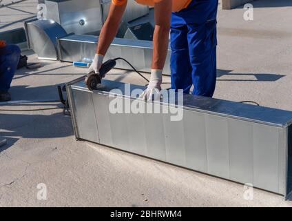 Il tecnico HVAC sta lavorando su un tetto di nuovo edificio industriale. Vista ravvicinata del giovane tecnico che ripara un condotto d'aria con la smerigliatrice angolare Foto Stock