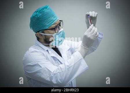 Giovane medico caucasico che guarda il campione di sangue in una provetta con maschera chirurgica e guanti Foto Stock