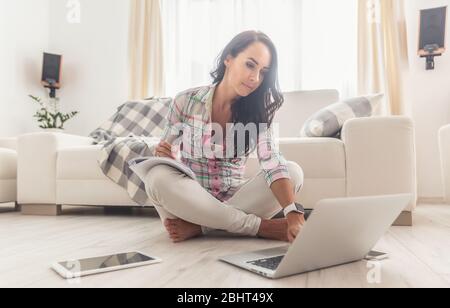 Giovane donna che studia da un notebook sul allagamento, seduta a gambe incrociate con un blocco note sul ginocchio in appartamento. Concetto di ufficio domestico. Foto Stock