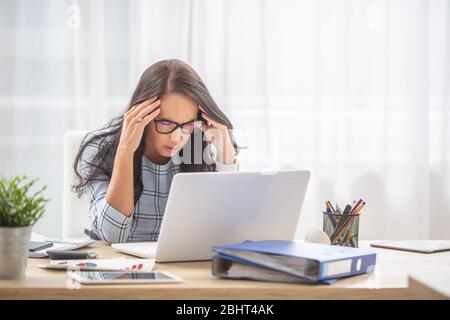 Donna che ha una testa di emigrene che tiene nelle sue mani con gli occhi chiusi alla scrivania, davanti al computer. Foto Stock