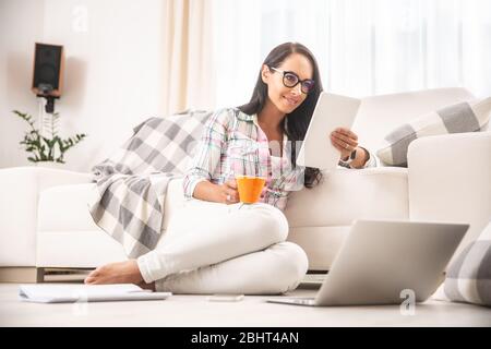 Buona ragazza che guarda in bicchieri leggere una tavoletta e bere caffè appoggiata su un divano e seduta su un pavimento riscaldato. Concetto di ufficio domestico. Foto Stock