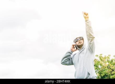 Un giovane UN giovane uomo con occhiali da sole blu, felpa grigia e barba che parla su un telefono cellulare guardando in su Foto Stock