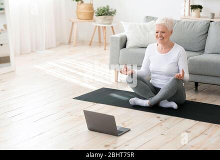 Yoga online. Donna senior rilassata che medita davanti al notebook a casa Foto Stock