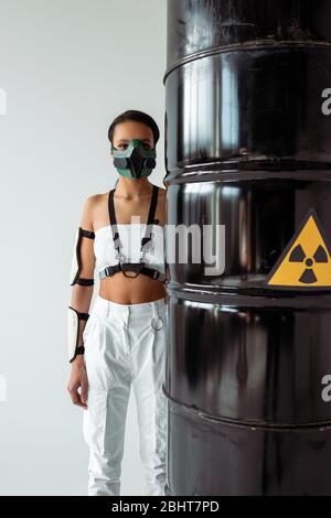 donna futuristica african american in maschera di sicurezza vicino ai barili di scorie radioattive isolati su bianco Foto Stock