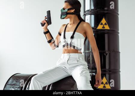 donna futuristica african american in maschera di sicurezza con pistola vicino ai barili di scarico radioattivo isolati su bianco Foto Stock