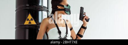 donna futuristica african american in maschera di sicurezza con pistola vicino barili di rifiuti radioattivi isolati su bianco, colpo panoramico Foto Stock