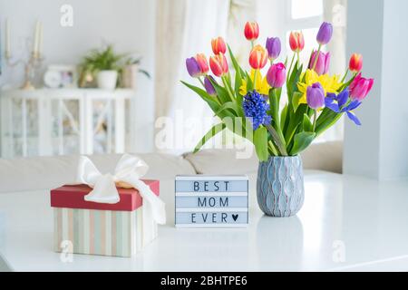 Sfondo della Giornata della mamma felice. Fiori di primavera colorati bouquet in vaso, scatola regalo con nastro satinato e scatola luminosa con parole miglior mamma mai su bianco marb Foto Stock