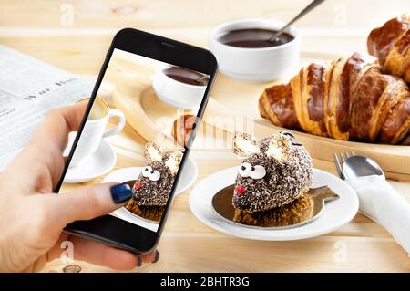 Donna mano prende telefono foto di cibo. Colazione continentale con croissant, torta fatta a base di mouse e caffè su fondo di legno. Smartph Foto Stock