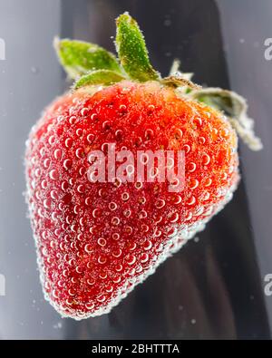 Una fragola rossa matura immersa in un liquido limpido con molte bolle. Riflessioni astratte sul contenitore di vetro. Foto Stock