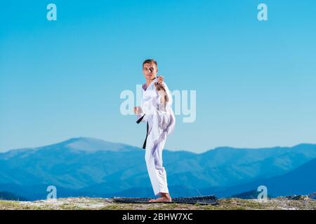 Bionda atleta di karate non kata sulla cima di una montagna mentre si esegue una linea di calci, pugni e blocchi sulla cima di una montagna in una giornata di sole. Foto Stock