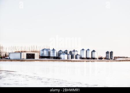 Acqua che sale in un campo agricolo minacciando un cantiere con bidoni di grano di acciaio e deposito di attrezzature capannone in un paesaggio rurale primaverile Foto Stock