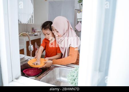 Aiuto mano. Cute Little Girl aiuta la sua madre musulmana nel lavare i piatti alla cucina di famiglia Foto Stock