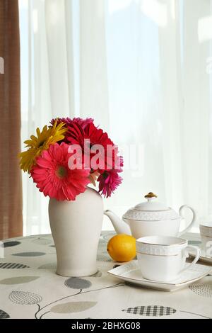 Gerbera colorata in vaso con teiera, tazze e limoni sul tavolo su sfondo tende Foto Stock