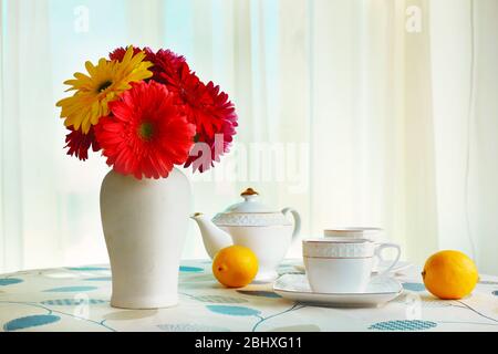 Gerbera colorata in vaso con teiera, tazze e limoni sul tavolo su sfondo tende Foto Stock