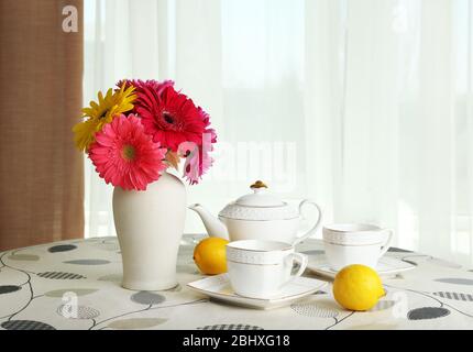 Gerbera colorata in vaso con teiera, tazze e limoni sul tavolo su sfondo tende Foto Stock