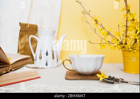 tazza bianca in porcellana con cucchiaio d'argento con granuli di tè e forsitia gialla su tavolo chiaro, sullo sfondo di una teiera bianca, vecchi libri a. Foto Stock