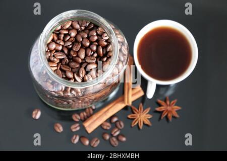 Caffè con spezie, chicchi di caffè tostati scuri in vaso di vetro, anice stellato, bastoncini di cannella e tazza bianca su tavolo di vetro scuro Foto Stock