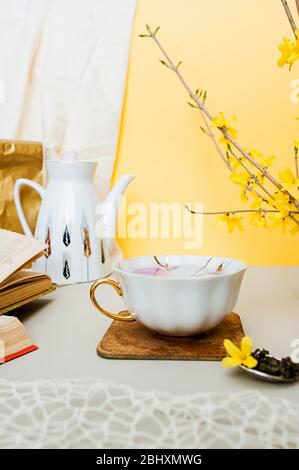 tazza bianca in porcellana con cucchiaio d'argento con granuli di tè e forsitia gialla su tavolo chiaro, sullo sfondo di una teiera bianca, vecchi libri a. Foto Stock