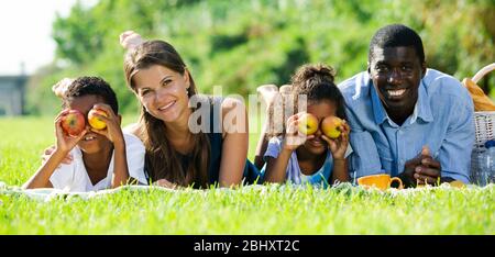 Allegro razza mista coppia con bambini preteen allegramente trascorrere il tempo in picnic, giacente insieme sul prato verde Foto Stock