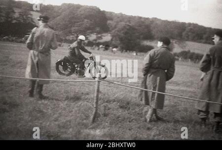Anni '1950, storico, uno scrambler sulla sua motocicletta dell'epoca che corre lungo una collina erbosa, guardato da tre funzionari in lunghi camici all'interno della zona cordata, Inghilterra, Regno Unito. Foto Stock
