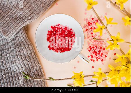 festa primaverile del tè. piccola tazza bianca riempita con stelle rosa e rosse del faretto e piattino su uno sfondo chiaro con i fiori gialli della forsithia della molla. Piatto Foto Stock