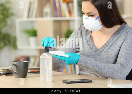 Donna con maschera protettiva e guanti applica igienizzante per disinfettare lo smartphone a causa di 19 anni seduto su una scrivania a casa Foto Stock