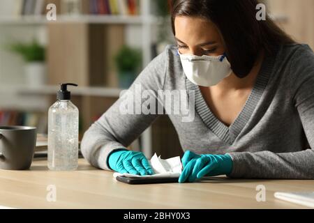 Donna con guanti e maschera protettiva che disinfettano lo smartphone da coronavirus seduto su una scrivania di notte a casa Foto Stock