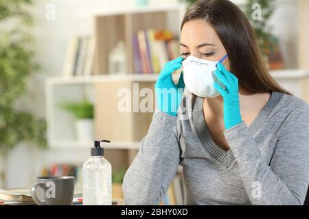 Donna che mette maschera protettiva due coronavirus con guanti seduti su una scrivania a casa Foto Stock