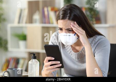 Ragazza preoccupata con maschera protettiva che legge le notizie difettose di coronavirus sullo smart phone che vetta su una scrivania a casa Foto Stock