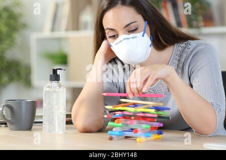 Donna pigra annoiato con maschera protettiva in quarantena confinamento dovuto coronavirus perdere tempo giocando con matite a casa Foto Stock