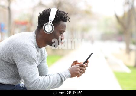 Profilo di un uomo nero serio con cuffie che ascoltano musica su smartphone seduto su un panca di parco Foto Stock