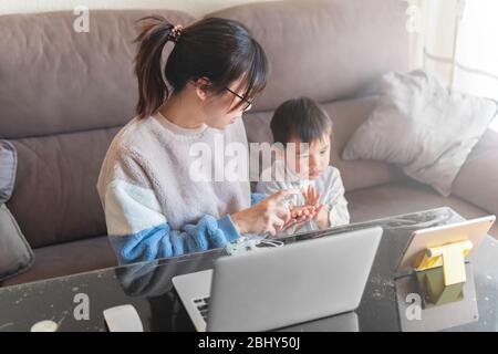 Giovane madre asiatica che lavora su un moderno notebook da casa con il bambino che guarda i cartoni animati su un tablet durante il blocco pandemico del coronavirus Foto Stock