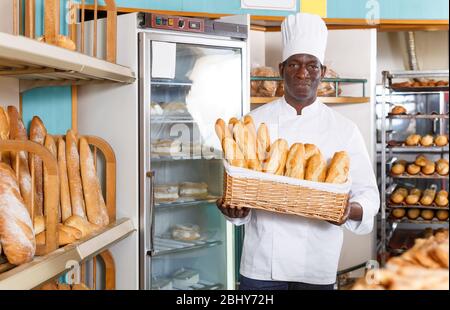 Chef di panetteria afroamericana in uniforme bianca che tiene un cesto di vimini con baguette appena sfornate al bakeshop Foto Stock