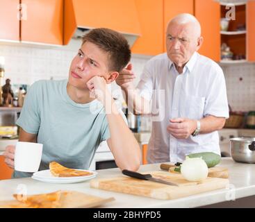 Ragazzo accigliato seduto al tavolo da cucina, ascoltando rimproverare dal nonno scontento Foto Stock