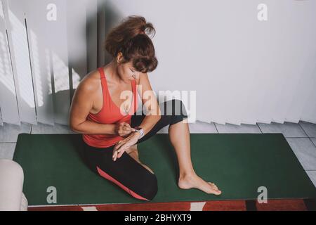 Atletica bella donna di mezza età in stretto abbigliamento sportivo, seduta su tappetino yoga dopo gli allenamenti, controllo degli indicatori di salute del corpo su smart watch, fitness tracker. Attività sportiva al coperto Foto Stock