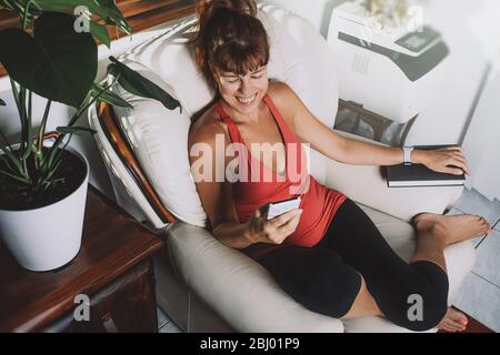 Affascinante donna di mezza età che guarda smartphone e sorridente, seduta con un libro seduto su una poltrona in un soggiorno. Foto Stock