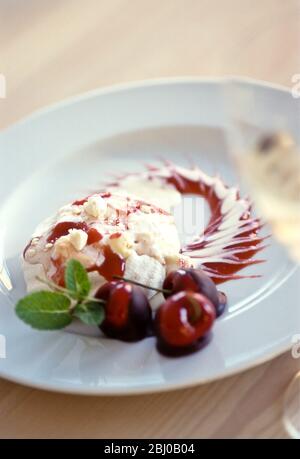 Spettacolare, dessert di meringa con gelato alla vaniglia, coulis di ciliegia e ciliegie fresche immerse in cioccolato fondente su piatto bianco guarnito con menta Foto Stock